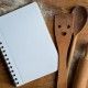 Open recipe book with wooden spoon, rolling pan and spatula  on wooden background, selective focus. Cooking course poster background - layout with free text space.