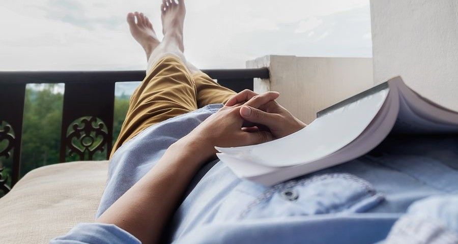 Young man reading a book lying on soft mattress in relaxing bed at terrace with green nature view. Fresh air in the morning of weekend or free day. Relax or education background idea. Selective focus.