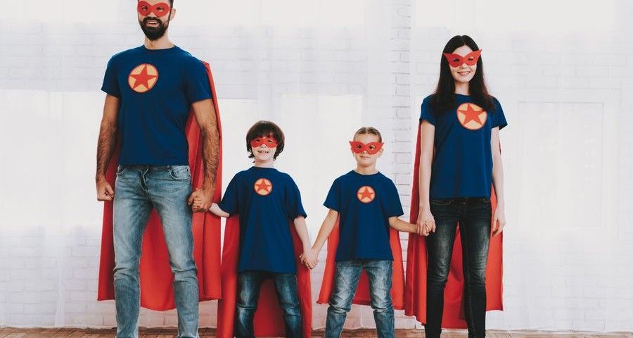 Young Family In Superhero Suits. Posing Concept. Masks And Raincoats. Bright Room. Resting Together. Save The World. Get Ready. Hand In Hand. Kids With A Parents. Active Lifestyle.