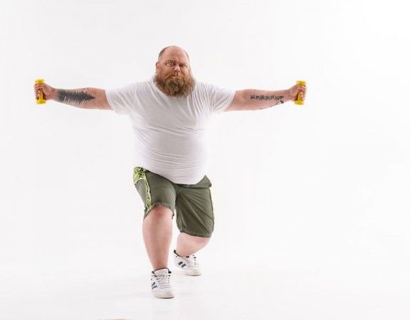 Fat man is lifting dumbbells with efforts. He is standing and stretching arms sideways. Isolated